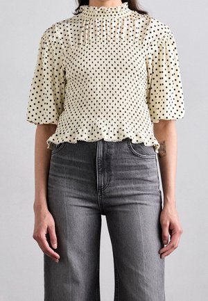 Mujer que lleva una blusa crema ajustada con lunares negros y mangas abullonadas, combinada con vaqueros grises de talle alto.