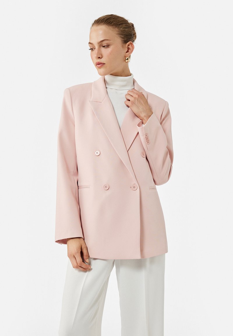 Lys rosa dobbeltbrystet blazer med store knapper, spidse revers og sidelommer, parret med en hvid turtleneck og neutrale bukser.