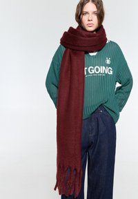 Burgundy scarf i mjukt material, har en tjock, texturerad design med fransade ändar, kombinerad med en grön ribbad tröja och denimbyxor.