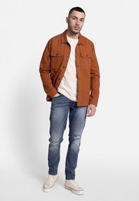 Roostevärvi nööbitav jakk kahe rinnataskuga, kantud kreemja t-särgi peal, sobitatud helesiniste kulunud teksade ja valgete tossudega.