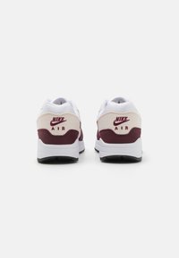 Nike Air tenisice, s bijelom i tamnocrvenom bojom, glatkom gornjom površinom i prepoznatljivim swoosh logom na peti. Teksturirane potplate.