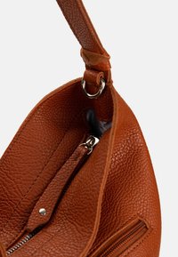 Sac à main en cuir marron texturé avec une poche zippée visible et un détail d'anneau en métal sur la bandoulière, sur un fond uni.