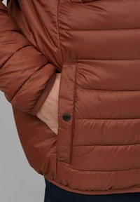 Bruine pufferjas met horizontale stiksels, voorzien van een zwarte drukknoop en rib gebreide manchetten. Glad, waterafstotend materiaal.