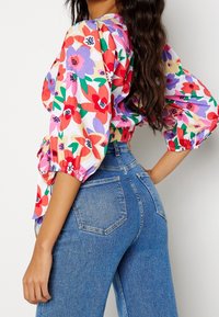 Blouse fleurie colorée avec des manches bouffantes, associée à un jean en denim taille haute. La blouse présente des motifs floraux rouges, violets et verts.