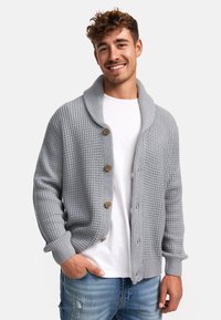 Grauer gestrickter Cardigan mit weitem Kragen, knöpfbarem Vorderteil und strukturiertem Muster, getragen über einem weißen T-Shirt und kombiniert mit blauen Jeans.
