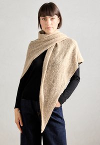 Châle en laine beige tricoté, présentant un design texturé et superposé avec une coupe angulaire. Porté par-dessus un haut noir et un jean bleu.