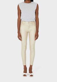 Henkilö, jolla on yllä ilmanhiha harmaa paita, korkeavyötäröiset beige skinny-housut ja valkoiset avokkaat, seisoo vaaleaa taustaa vasten.