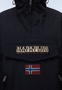 Marineblå vindjakke med høj krave, prydet med et stort patch-logo og et flagmærke for Norge. Materialet ser holdbart ud med en glat tekstur.