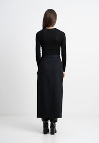 Kobieta o długich brązowych włosach, ubrana w czarną bluzkę z długim rękawem, czarną spódnicę midi i czarne botki na obcasie, stoi tyłem do przodu.