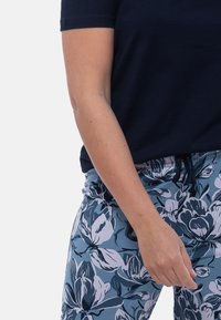 Henkilö, jolla on päällään tummansininen lyhythihainen paita ja sinivalkoisen kukkakuosiset housut, joissa on naru, käsi rentoutuneena sivulla.