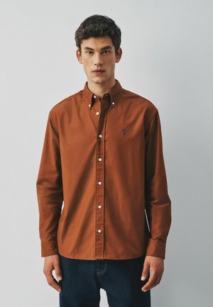 Young man with short dark hair wearing a rust-colored button-up shirt and dark jeans against a plain light background.