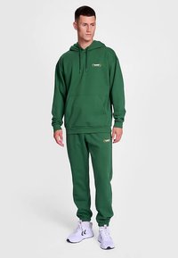Grön hoodie med justerbara dragsnören, framficka och logotyp. Matchas med joggingbyxor med resår i benslut och sidodetaljer.