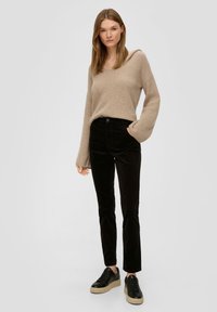Femme portant un pull en tricot beige, un pantalon noir et des baskets à plateforme noires, se tenant avec une main dans la poche sur fond uni.