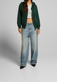 Felpa verde con zip sopra un top grigio corto, abbinata a jeans a gamba larga azzurri. Tessuto morbido con una vestibilità casual, dettagli minimi.