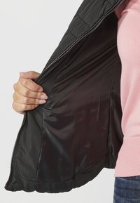 Črn prešit jakna z sijočim notranjim delom, ki prikazuje zadrgo ob sredini spredaj. Zunanji material se zdi mehak in gladek.
