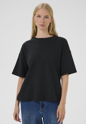 Femeie cu păr blond lung, purtând o bluză neagră largă cu mâneci scurte și blugi albaștri, stând în fața unui fundal simplu, deschis la culoare.