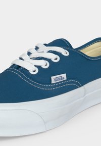 Baskets Vans bleues avec lacets blancs, semelle blanche, coutures visibles et petite étiquette avec logo Vans sur le côté.