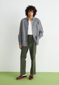 Geruit grijs en wit overhemd, witte t-shirt eronder, donkergroene broek, bruine en beige schoenen, staand op een groene ondergrond met een neutrale achtergrond.