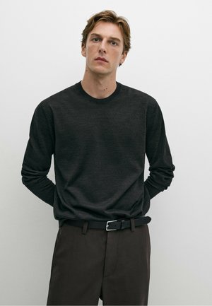 Junger Mann mit hellbraunen Haaren, der einen dunklen Langarmpullover und dunkle Hosen mit schwarzem Gürtel trägt, steht vor einem schlichten hellen Hintergrund.