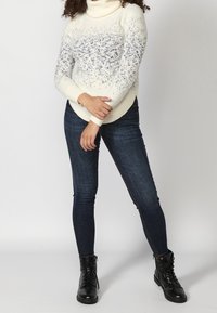 Maglione a collo alto color crema con motivo a macchie blu, abbinato a jeans skinny scuri e stivaletti neri con lacci e dettagli testurizzati.