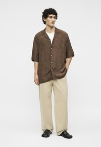 Νέος άνδρας με σγουρά μαλλιά, που φορά καφέ πουκάμισο με μοτίβο και κοντά μανίκια, μπεζ φαρδιά παντελόνια και μαύρα παπούτσια, στέκεται με το ένα χέρι στην τσέπη.