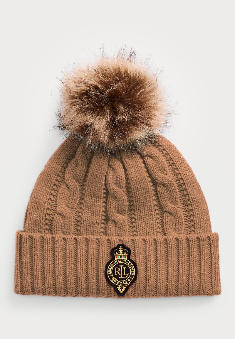 Brūns adīts beanie cepure ar kabeli rakstu, ar mākslīgā pūka pom-pomu un aplītu melnu logo uzšuvumu ar zelta akcentiem priekšpusē.