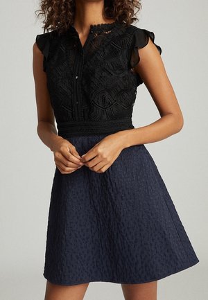 Femme portant une robe noire en dentelle sans manches avec des épaules volantées et une jupe texturée bleu marine, les mains jointes à la taille.