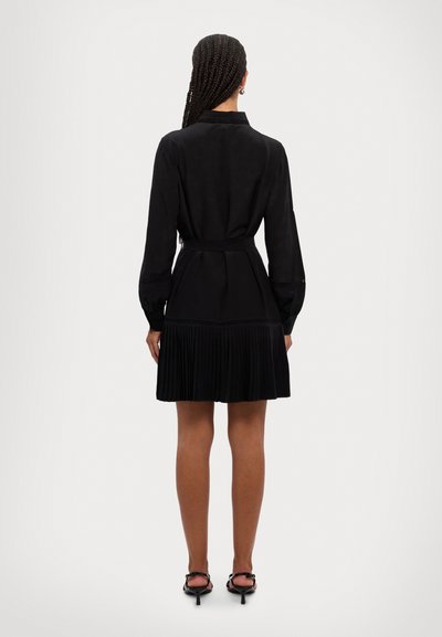 Vestido negro plisado con mangas largas, cuello con solapa y un lazo en la cintura. Textura suave con una falda acampanada que llega hasta mitad del muslo.