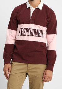 Rugbyshirt met bordeauxrode en roze strepen en een kraag, met de tekst "ABERCROMBIE" in donkere letters over de borst.