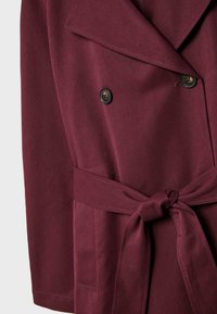 Burgundy trench coat dengan lapel lebar, kancing ganda, dan sabuk kain yang diikat di pinggang.