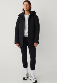 Jongeman in een zwarte hooded jacket, grijze sweatshirt, zwarte legging, witte sokken en witte sneakers die tegen een effen lichte achtergrond staat.