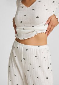 Conjunto de pijama blanco que presenta una blusa texturizada con un escote en forma de onda y bordados florales, acompañado de pantalones a juego que muestran patrones similares.