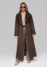 Abrigo largo marrón a rayas con amplias solapas, combinado con un suéter y pantalones beige. Los accesorios incluyen gafas de sol y pendientes grandes.