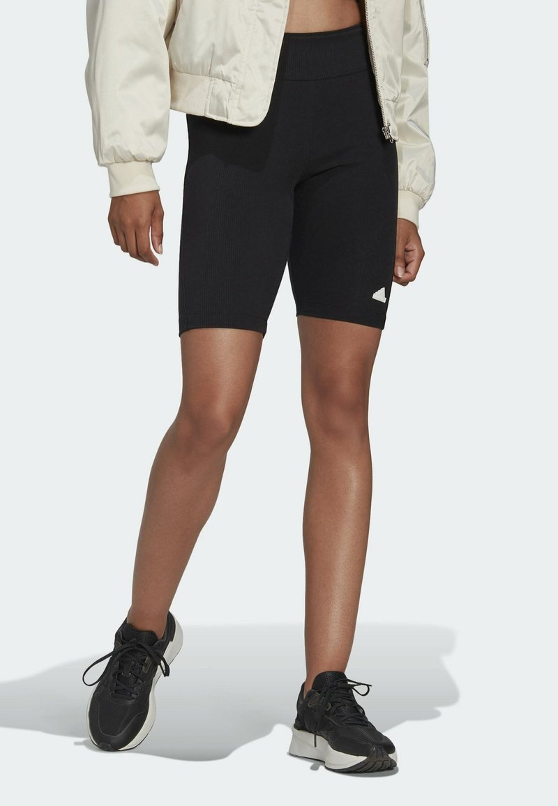 Svarta ribbade cykelshorts med hög midja och en liten vit logotypdetalj till vänster. Bärs med en ljusbeige bomberjacka och svarta sneakers.
