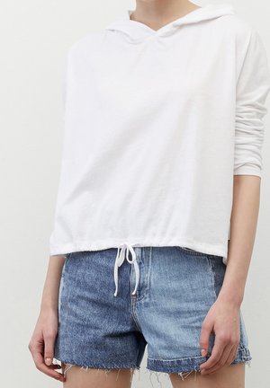 Person trägt ein weißes, langärmliges, kurzes Hoodie mit Kordelzug am Taillenbund, kombiniert mit zweifarbigen Jeansshorts mit ausgefransten Säumen.