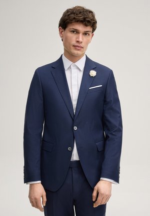 Giovane uomo con indosso un abito blu navy, una camicia bianca, una spilla a forma di fiore bianco all'occhiello e un fazzoletto da taschino bianco abbinato, in piedi di fronte a uno sfondo semplice.