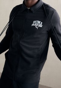 Chaqueta deportiva negra con cremallera y cuello, que presenta el logo blanco de "PUMA sports club" y una tela lisa y texturizada.