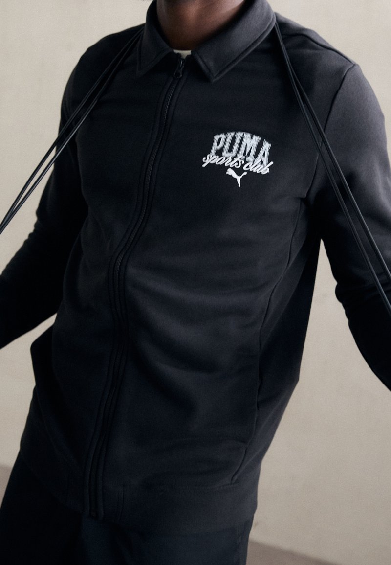 Chaqueta deportiva negra con cremallera y cuello, que presenta el logo blanco de "PUMA sports club" y una tela lisa y texturizada.