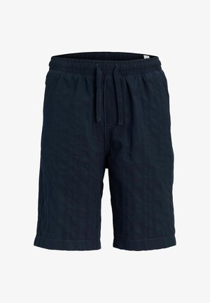 Niño con cabello rizado lleva una camiseta blanca de manga corta, pantalones cortos azul marino, calcetines crema y zapatos marrones con cordones, de pie frente a un fondo liso.