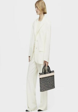 Bolso de portátil con un diseño negro y beige, que presenta un patrón oscuro texturizado, asas negras y una franja de acento crema. Llevado por un modelo con un traje blanco.