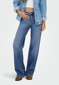 Jean large bleu à taille haute, en denim. Associé à un crop top blanc et une veste en denim bleu clair.