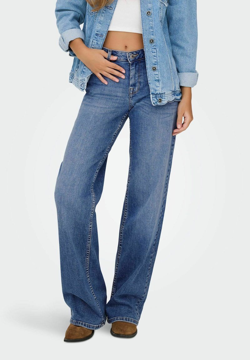 Jean large bleu à taille haute, en denim. Associé à un crop top blanc et une veste en denim bleu clair.