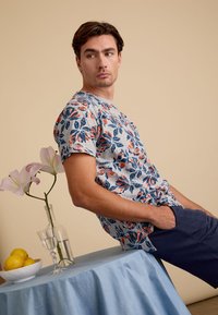 Korte mouwen T-shirt in lichtgrijs met een blauw en oranje bloemenpatroon, gecombineerd met donkerblauwe shorts. Tegen een beige achtergrond.