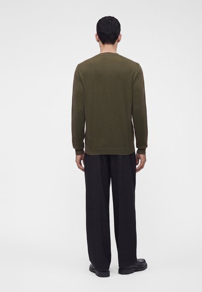 Homme aux cheveux courts foncés portant un pull vert olive et un pantalon noir ample, debout face à l’arrière sur un fond clair uni.