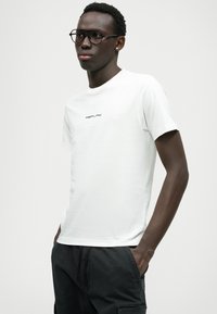 Biała bawełniana koszulka z okrągłym dekoltem, z czarnym logo "REPLAY" na piersi. Model nosi czarne okulary i szorty cargo.