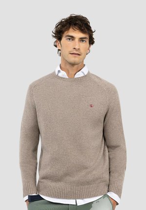 Hombre con suéter de punto beige sobre camisa blanca con las manos en los bolsillos contra un fondo liso gris claro.