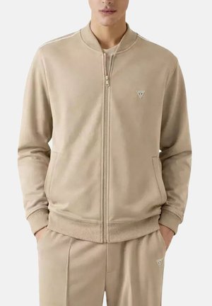 Man met beige jas met ritssluiting en bijpassende broek, handen in de zakken, staand tegen een effen achtergrond.