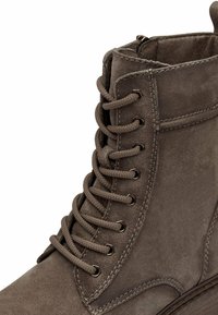 Ruskea mokkanahkasaapas nilkkapituisena, jossa on metalliset rei'itykset ja pyöreät nauhat, teksturoitu pinta sekä ommellut yksityiskohdat sivuilla ja pohjassa.