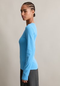 Langsleeve, getailleerde top van zachte, ribbelstof; heldere blauwe kleur; ronde halslijn; gedragen met een donkergrijze rok. Glad oppervlak.