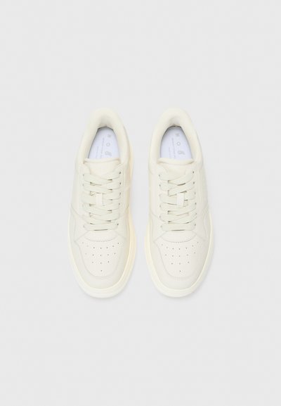 Des baskets blanches en cuir présentent des perforations sur les orteils, des lacets plats et une semelle en caoutchouc de couleur crème discrète. Design minimaliste sans motifs.
